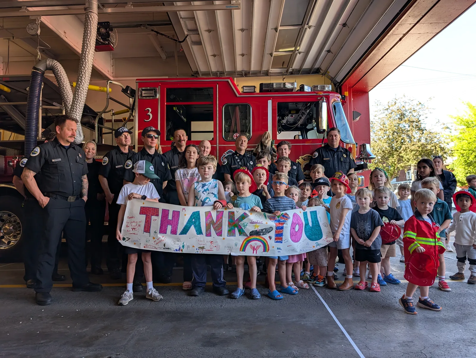 Dozens of small children and families pose in front of Engine 3 inside Station 3, alongside a row of Portland firefighters. The kids hold a hand-decorated paper banner reading 'THANK YOU', and several wear plastic red fire helmets.